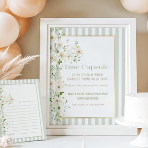 以下が含まれることがあります： 「タイムカプセル」の文字と花の装飾が施された白いフレームの看板と、お揃いのメモ帳。看板は白い花と緑のストライプで飾られています。台座に乗った白いケーキと背景の風船。