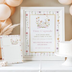 以下が含まれることがあります： 「タイムカプセル」の文字と花柄のデザインが施された額入りの看板とメモ帳。 看板は白いフレームに金色の縁取りとピンクのストライプ。 メモ帳は罫線入り。 背景には風船とケーキがあります。