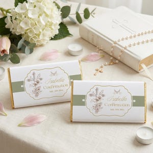 May include: Two white chocolate bars with gold foil edges, featuring a cross and floral design, and the text "Isabella Confirmation." A white Bible, rosary, and candles are also present, suggesting a religious theme.