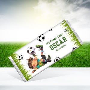 May include: A white chocolate bar wrapper with a cartoon illustration of a boy playing soccer. The wrapper has green grass and soccer balls on it. The text on the wrapper says "It's Game Time OSCAR 03-25-20xx".