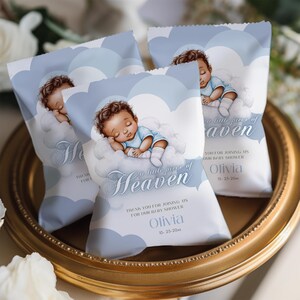 May include: Baby shower favor bags with a blue and white cloud design. Each bag features an illustration of a sleeping baby and the text "a little piece of Heaven". The bags are on a gold-rimmed tray.