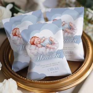 May include: Baby shower favour bags with a light blue cloud design and a sleeping baby illustration. The bags have the text "a little piece of Heaven" and "Olivia 10-25-20xx". The bags are on a gold-coloured tray.