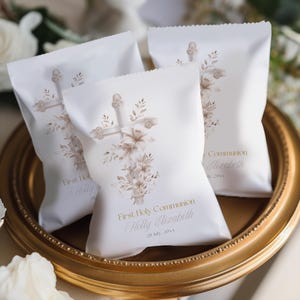 Könnte beinhalten: Drei weiße Gastgeschenktüten mit einem Kreuz- und Blumendesign in Beige- und Goldtönen. Auf den Tüten steht "First Holy Communion" sowie ein Name und ein Datum. Die Tüten sind auf einem goldfarbenen Tablett angeordnet.