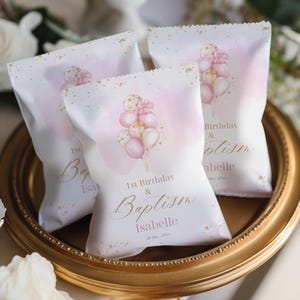 Könnte beinhalten: Drei weiße Gastgeschenktüten mit rosa Aquarellakzenten und goldenem Konfetti. Jede Tüte zeigt eine Ansammlung von rosa und weißen Luftballons mit einem Kreuz und dem Text "1. Geburtstag & Taufe Isabelle".