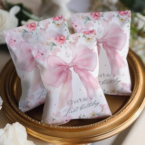 May include: Three white party favor bags with a pink bow and floral design. The bags have the text "Charlotie's First Birthday" printed on them. The bags are sitting on a gold-colored tray.