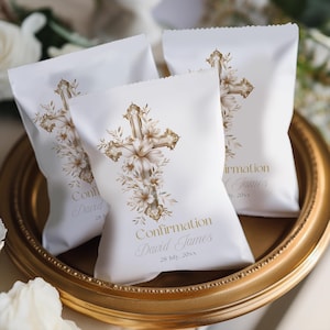 May include: White favor bags with a gold cross design and floral accents. The bags are personalized with the name "David James" and the date "28 July, 20xx" in gold script. The bags are arranged on a gold-colored tray.