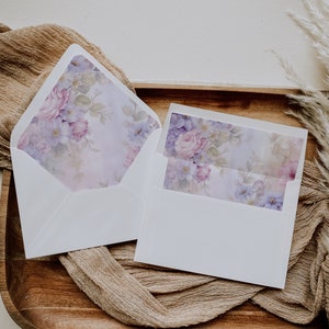 May include: Two white envelopes with a floral design on the front. The envelopes are on a brown surface with a beige fabric.