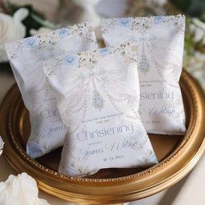 Könnte beinhalten: Drei weiße rechteckige Geschenktüten mit blau-weißem Blumenmuster, einer großen weißen Schleife, Perlenschnüren und dem Text: Thank you for joining us for the christening of our son James Wilson 10-23-20xx.