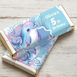 Op de afbeelding: Twee chocoladereepverpakkingen met een blauw en roze aquatisch ontwerp. Een cartoon dolfijn met paarse en blauwe accenten is te zien, samen met de tekst "Olivia's 5th Birthday". De verpakkingen hebben gouden folieranden en zijn geplaatst tegen een lichte houten achtergrond.