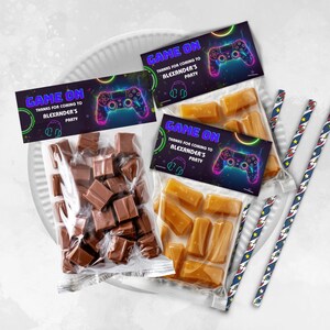 May include: Two clear cellophane bags filled with candy. One bag has chocolate squares and the other has caramel candies. Each bag has a colorful gamer-themed thank you tag that says "Game On" with a neon green, blue, and purple video game controller graphic. The text on the tag says "Thanks for coming to Alexander's Party."