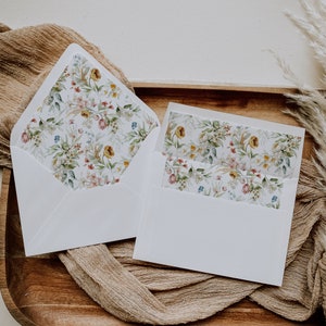 Könnte beinhalten: Zwei weiße Umschläge mit floralen Mustern. Die Umschläge liegen auf einer braunen Holzoberfläche mit einem beigefarbenen Stoff darunter.