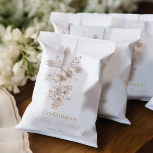 May include: White favor bags with a cross and floral design. The bags have the word "Confirmation" and a name and date printed in gold. The bags are sealed at the top and are sitting on a wooden surface.