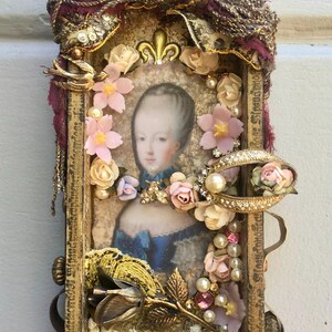 May include: A vintage-style framed portrait featuring a woman in a blue dress, surrounded by pink and cream-colored flowers, pearls, and gold embellishments. The frame is adorned with gold accents, a small crown, and a decorative brooch.