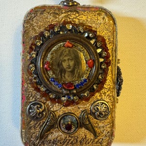 Puede incluir: Caja rectangular ornamentada con superficie dorada texturizada. Un marco circular central presenta un retrato, rodeado de joyas rojas, azules y transparentes. Adornos adicionales incluyen alas, detalles metálicos decorativos y un pequeño asa en la parte superior.