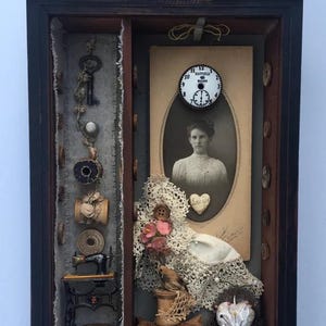 Könnte beinhalten: Eine dunkle Holzschatulle mit verschiedenen Vintage-Artikeln. Enthält ein Schwarz-Weiß-Foto, eine kleine Uhr, Nähutensilien und eine Miniatur-Nähmaschine. Die Schatulle hat eine dekorative Oberseite und weiße Knöpfe.