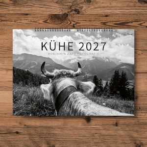 Könnte beinhalten: Schwarz-Weiß-Kalender für 2027 mit einer Kuh in einer Berglandschaft. Der Kalender hat eine Spiralbindung und den Text "KÜHE 2027" und "BENJAMIN ZAPF FOTOGRAFIE". Das Bild ist vor einem Holzuntergrund platziert.