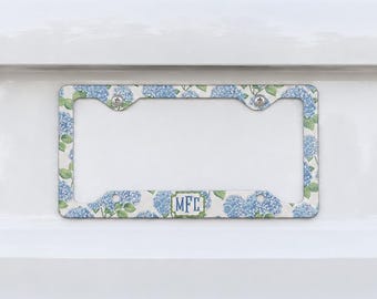 May include: A rectangular license plate frame featuring a repeating pattern of blue hydrangea flowers and green leaves on a white background. The bottom center of the frame displays a rectangular white monogram label with the black letters MFC.