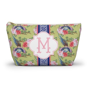 May include: A zippered cosmetic bag with a floral and bird pattern in shades of pink, blue, and green. The bag features a white and pink monogram "M" on the front, with a navy blue and white stripe down the center.