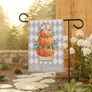 Peut inclure: Un drapeau de jardin avec une pile de citrouilles orange, des feuilles vertes et un nœud blanc. Le drapeau présente un motif de losanges gris et blancs et le texte "The Carters". Il est affiché sur un support en métal noir.