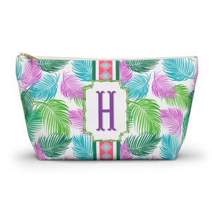 May include: A white zippered pouch with a tropical palm leaf pattern in shades of green, teal, and purple. The pouch features a central design with the letter "H" in purple, framed by a bamboo-style border and a pink and teal stripe.