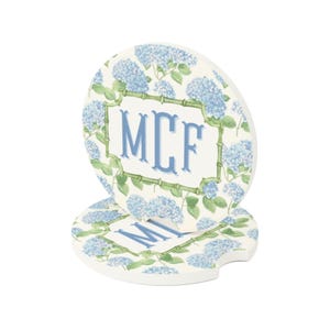 May include: Two white ceramic car coasters with a floral hydrangea design. The coasters feature a light blue floral pattern and a green bamboo-style border. The center of the coaster displays the initials "MCF" in blue.