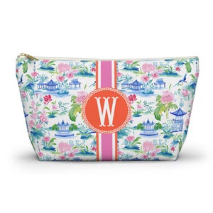 Op de afbeelding: Een witte make-uptas met een kleurrijk bloemen- en pagodepatroon. De tas heeft een roze en oranje streep in het midden met de letter "W" in een cirkel. De tas heeft een gouden rits.
