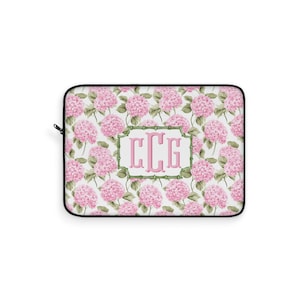May include: Laptop sleeve featuring a floral pattern of pink hydrangeas on a white background. The center has a monogram in a bamboo-style frame. The sleeve has a black border and a zipper.