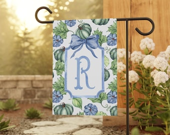 Könnte beinhalten: Eine Gartenflagge mit einem Aquarelldesign mit blauen Kürbissen, Blättern und Blumen. Eine blaue Schleife und ein gerahmter Buchstabe "R" sind auf der Flagge zentriert. Die Flagge wird auf einem schwarzen Metallständer in einem Garten gezeigt.