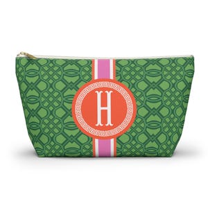 Pode incluir: Bolsa de cosméticos verde com um padrão geométrico. A bolsa apresenta uma faixa vertical rosa e branca e um emblema circular laranja com a letra "H" branca. O zíper é dourado.