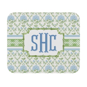 Puede incluir: Alfombrilla de ratón rectangular con un estampado floral de hortensias azules y hojas verdes. El centro presenta las letras azules "SHC" dentro de un marco verde estilo bambú, sobre un fondo blanco.