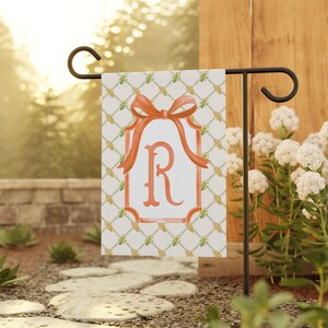 Peut inclure: Un drapeau de jardin avec un nœud orange et la lettre "R" dans un cadre rectangulaire. Le drapeau a un fond crème avec un motif de treillis et est affiché sur un support en métal noir. Décoration de jardin.