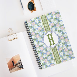 May include: A floral patterned spiral-bound notebook featuring white and pink flowers on a light blue backdrop. A green and white Greek key design stripe runs down the centre. The letter "H" is in a green font within a white and light blue scalloped frame.