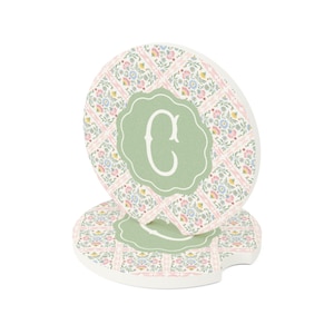 May include: Two round car coasters with a floral pattern in pink, blue, and yellow. Each coaster has a light green center with the letter "C" in white. The coasters are designed to fit in a car's cup holder.