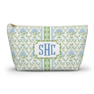 May include: A white cosmetic bag with a floral pattern of blue hydrangeas and green leaves. The bag features a central panel with the initials "SHC" in blue, framed by a green border. A gold zipper secures the top.