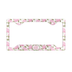 May include: White license plate frame with a pink floral hydrangea pattern. The frame has a rectangular shape with rounded corners and a cutout for the license plate. The text "ARC" is in a pink box.