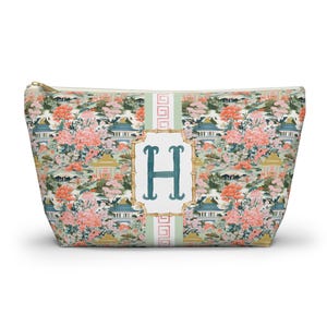 May include: A zippered cosmetic bag with a floral and pagoda pattern in shades of pink, green, and gold. The bag features a white rectangle with the teal letter "H" in the center.