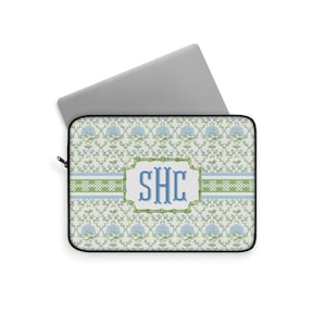 May include: Laptop sleeve with a floral pattern in light blue and green. The design includes a central rectangular frame with the initials "SHC" in blue. A silver laptop is partially visible.
