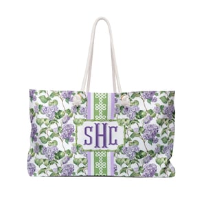 May include: Large tote bag with a white background and a floral pattern of purple hydrangeas and green leaves. The bag has a white rope handle and a central panel with the initials "SHC" in a purple frame.