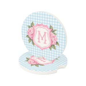May include: Two round car coasters featuring a light blue and white gingham pattern. Each coaster displays a pink shield-shaped crest with a central letter M, surrounded by pink hydrangea flowers and green leaves on a white background.