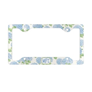 May include: White license plate frame with a pattern of blue hydrangea flowers and green leaves. The frame has a rectangular opening and the initials "MFC" in a blue box.