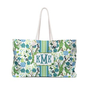 Puede incluir: Bolso tote grande con fondo blanco, con un estampado floral y de pájaros en tonos verdes y azules. El bolso tiene un panel central con las iniciales "KME" y asas de cuerda blanca.