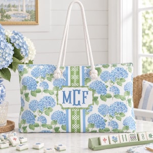 May include: A rectangular tote bag featuring a blue and white hydrangea floral pattern with a central vertical green and blue striped panel. The bag has white rope handles and a central bamboo-style frame containing the blue monogram letters MCF.