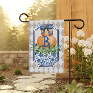 May include: A garden flag with a seasonal design. It shows orange pumpkins with a blue bow and the letter "B". Below the pumpkins is a blue and white floral vase filled with greenery. The background has a diamond pattern, perfect for a garden.