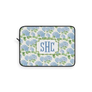 May include: A rectangular laptop sleeve featuring a floral pattern of blue hydrangeas and green leaves on a cream background. The center has a white rectangle with the letters "SHC" in blue. The sleeve has a black border and a black zipper.
