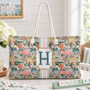 May include: A rectangular tote bag with white rope handles featuring a chinoiserie-inspired pattern of pagodas and floral motifs in shades of green, pink, and blue. A central white panel displays a large, dark green letter H in a bamboo-style frame.