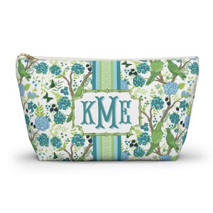 May include: A white cosmetic bag with a gold zipper, featuring a floral and bird pattern in shades of green and blue. The bag has a central panel with the letters "KME" in teal.