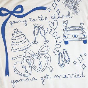 May include: White sweatshirt with blue line art. The design includes a bow, cake, champagne glasses, a car, hearts, shoes, and the phrases "going to the chapel" and "gonna get married." The date "07.02.22" is also included.