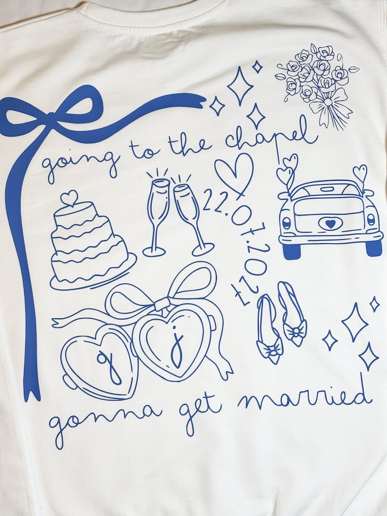 May include: White sweatshirt with blue line art. The design includes a bow, cake, champagne glasses, a car, hearts, shoes, and the phrases "going to the chapel" and "gonna get married." The date "07.02.22" is also included.