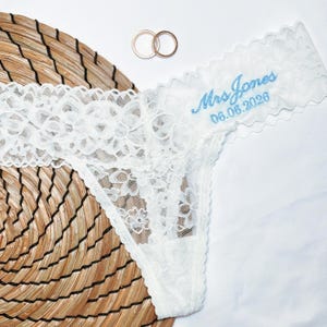 May include: White lace thong underwear with the embroidered text "Mrs Jones 06.06.2026" in light blue. Two gold wedding rings are visible in the background. The underwear is placed on a woven mat.