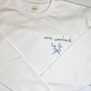May include: A white crew neck sweatshirt with the text "mrs woodcock to be" in navy blue script, along with a bow graphic. The sweatshirt has a small label near the neckline.